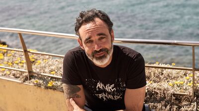 A man with salt-and-pepper facial hair and dark hair looks toward the camera with a gentle smile while sitting in a wheelchair near a coastal walkway with the sea in the background.