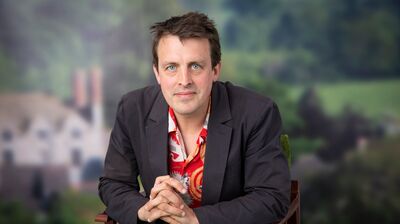 A man with blue eyes, a dark blazer, and a red patterned shirt sits with hands clasped, looking at the camera.