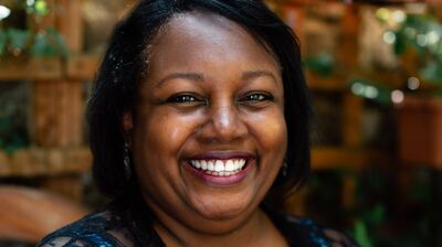 Portrait of author Malorie Blackman. She sits in a garden wearing a patterned blue top and a lace cover up.