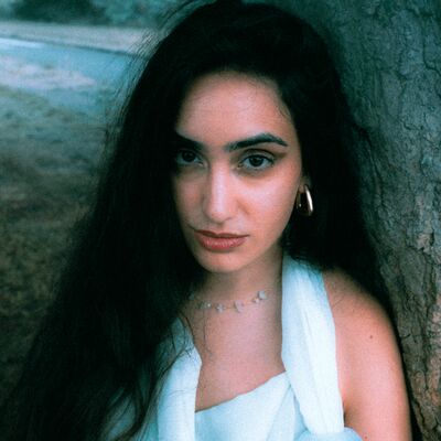 A woman with long dark hair and a light-colored top leans against a tree trunk in an outdoor setting.