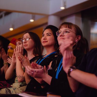 An audience sits in a row, looking up at a stage and clapping under warm lighting at a literary event or convention.
