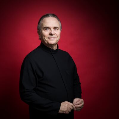 Portrait of a smiling man in a black high-collared shirt, posing against a solid deep red background.