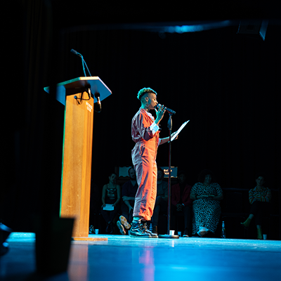 Someone in orange dungarees speaking on a stage in front of a microphone