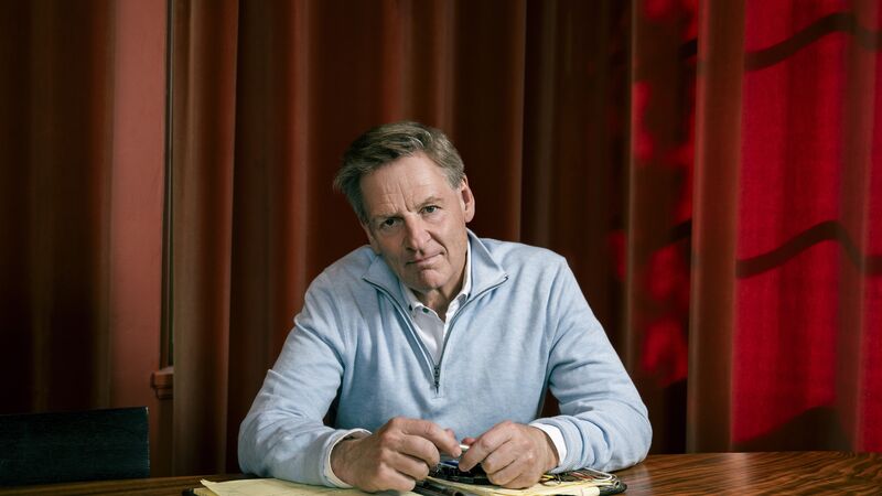 Man wearing a blue shirt sitting at a wooden table with a notebook against a background of red curtains.