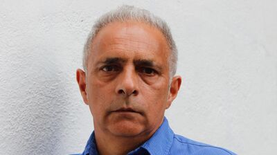 A man with grey hair wearing a blue shirt in front of a white wall