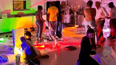 People participate in an interactive art workshop with papers under vibrant, colorful lighting.