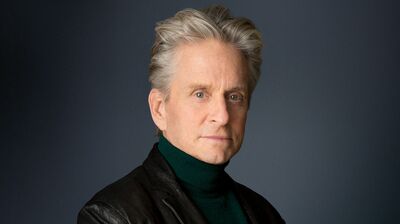 Michael Douglas with grey hair wearing a black leather jacket and a green turtleneck