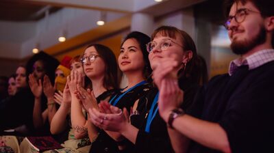 An audience sits in a row, looking up at a stage and clapping under warm lighting at a literary event or convention.
