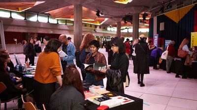 People browse books at tables in a large, wide brutalist hall with exposed concrete pillars and warm wooden ceiling lights.