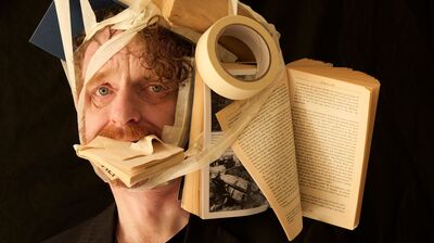 a man in a black blazer looking directly at the camera while a chaotic cluster of books and a roll of masking tape are bound tightly to his head and stuffed into his mouth using beige tape.