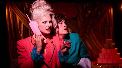 Two performers in bright 1980s jackets look off-stage with shocked expressions under dramatic red lighting.