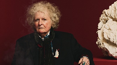 Woman with white curly hair wearing black standing in front of a red wall with her arm resting on a red plinth.