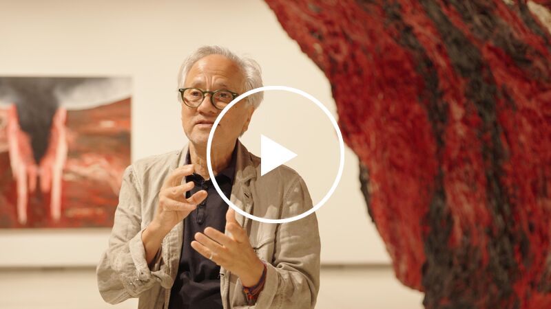 Anish Kapoor, an older man with white hair and glasses, stands in the Hayward Gallery between two of his artworks, a red sculptural piece and a painting hanging on a wall