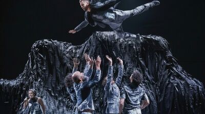 A dramatic dance performance with five performers in gray outfits tossing a sixth performer, also in gray, into the air above a large, dark, textured mound on a stage.