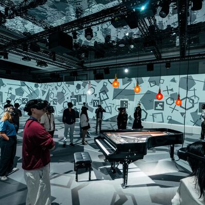 A wide shot of a spacious exhibition room where people wearing VR headsets walk around a grand piano beneath glowing orange bulbs, surrounded by geometric wall projections.