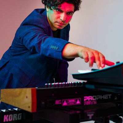A man in a blue suit reaches for a synthesizer keyboard, illuminated by dramatic pink and blue studio lighting.
