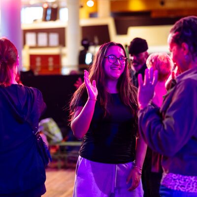 A smiling young woman with glasses waves to a group of people dancing under vibrant pink stage lights.