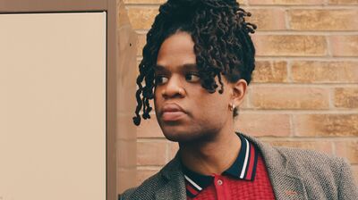 Author Cenac Rollins wearing a red polo shirt and tweed jacket, leaning against a locker in front of a brick wall