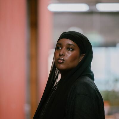 A three-quarter portrait of Momtaza Mehri looking upward, wearing a black hijab and matching black linen jacket.