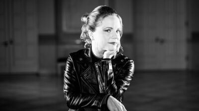 Black and white portrait of Julia Fischer, wearing a leather jacket, smiling and resting her chin on her hand