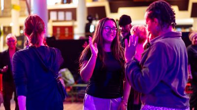 A smiling young woman with glasses waves to a group of people dancing under vibrant pink stage lights.