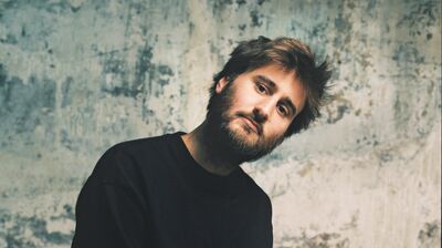 A bearded man in a black sweatshirt sits on a stool, tilting his head slightly against a distressed, gray concrete wall.
