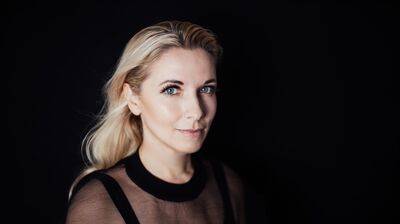 A studio portrait of a blonde woman wearing a black mesh top set against a solid dark backdrop.