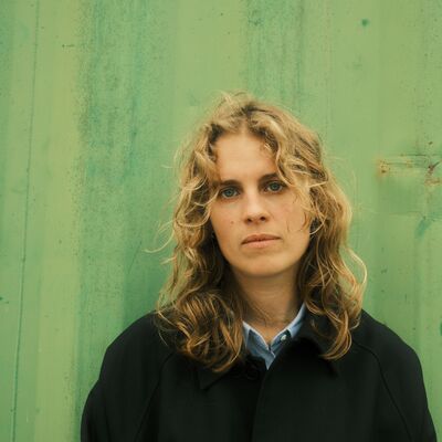A portrait of a woman with wavy blonde hair in a black coat standing against a textured green background.