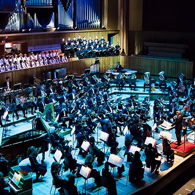 A wide shot of a full orchestra and choir performing on stage in a grand concert hall illuminated by cool blue light.