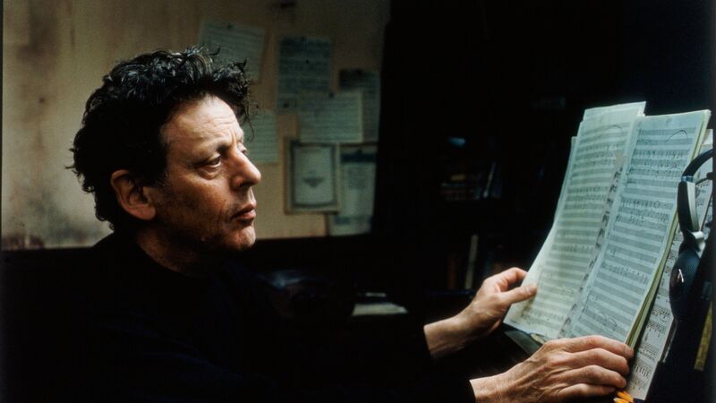 Composer Philip Glass sits at a piano, looking intently at sheet music in a dimly lit, workspace setting.