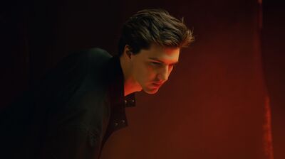 A close-up profile of a young man leaning forward, his face illuminated by a dramatic, warm orange glow against a dark backdrop.