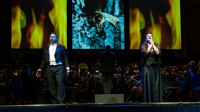 Two vocalists perform on stage before an orchestra, backed by large screens displaying flame and stone visuals.