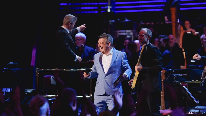 A performer in a light blue suit smiles on stage surrounded by musicians and an applauding audience.