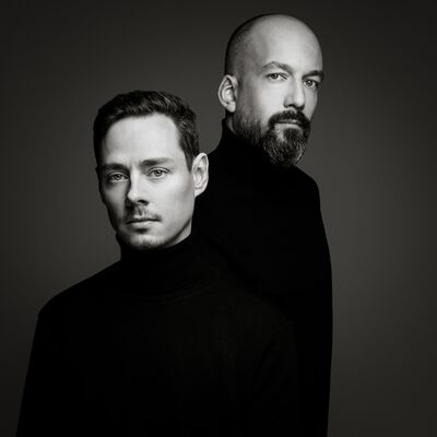A black-and-white studio portrait of two men in black turtlenecks standing staggered against a grey gradient background.