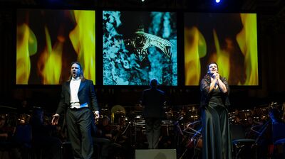 A man in a suit and woman in a black dress stand on stage singing with theatrical gestures.