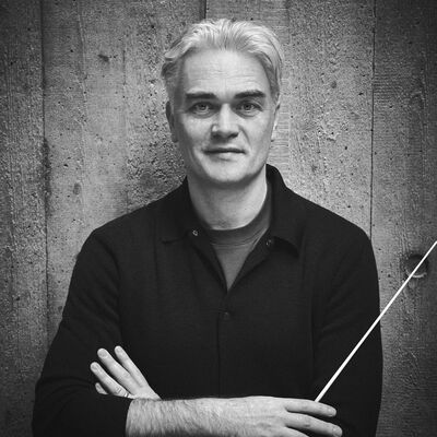Black and white portrait of conductor Ed Gardner, a man with short silver hair, wearing a black shirt with arms folded and holding a conductor's baton, looking at the camera against a stone wall.