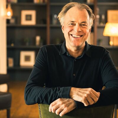 Thomas Hengelbrock, against a warm yellow background, sits and smiles at the camera