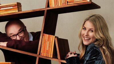 A man with glasses and a burgundy sweater peeks through an angular wooden bookshelf next to a smiling blonde woman.