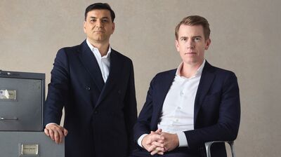 Two men in dark suits pose next to a gray filing cabinet and under a security camera against a neutral background.