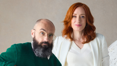 A man with a graying beard and glasses in a green sweater poses next to a woman with red hair in a white suit.