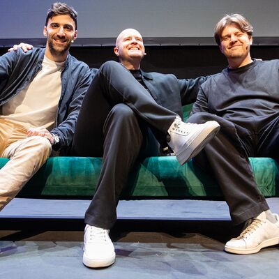 Three men sitting on a green sofa on an empty stage.