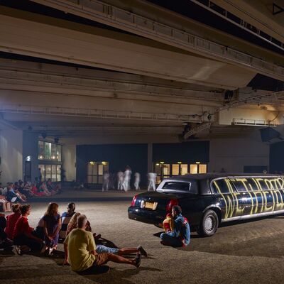 A crowd of people watching performers by a long, black limousine.