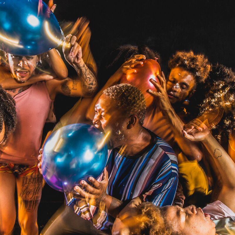 Performers grabbing each other whilst holding and popping balloons