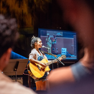 A woman on stage playing a guitar with a projector in the background.