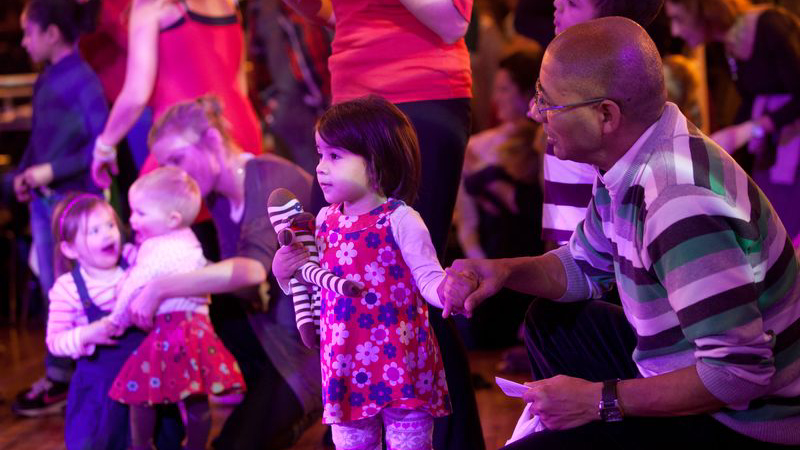 Father and Daughter at dance workshop. The father is crouching down looking at her and holding her hand. She clutches a teddy while watching something in the distance.