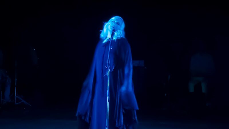 A neon blue Anohni standing in front of a mic draped in a dark blue dress.