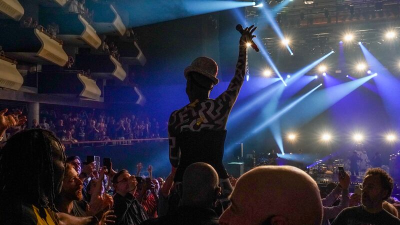 Grace Jones crowd-surfs in a packed Royal Festival Hall auditorium. She is lifted high and raising her hand in the air holding a microphone. You can see the back of her head wearing a sparkly bowler hat. Lights shine down and she has confetti over her.