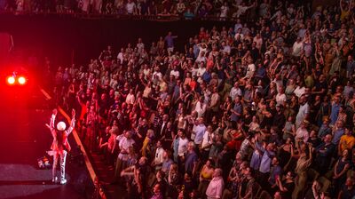 Nile Rodgers performing at his Meltdown Festival to a sold out crowd in the Royal Festival Hall. Red lights surround the auditorium. Nile Rodgers stands as the front of the stage with his back to the camera facing the audience with his hands raised. The audience is stood with their hands raised too.