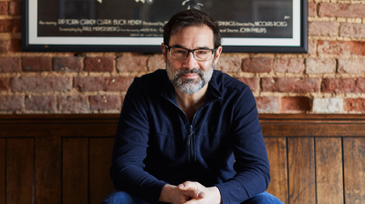 A man with glasses and a beard sits on a wooden bench, smiling in front of a brick wall and vintage poster