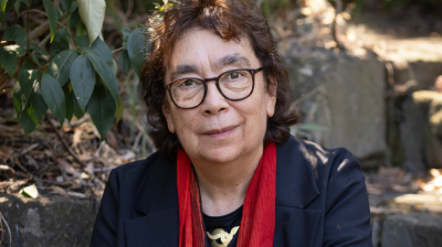 A woman with glasses and a red scarf sits on stone steps surrounded by greenery, smiling into camera.